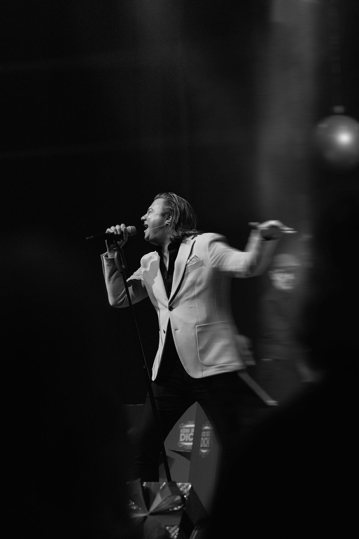 Marinho Peters zingt live in een witte colbertjas met microfoon in de hand, in zwart-wit onder podiumlicht